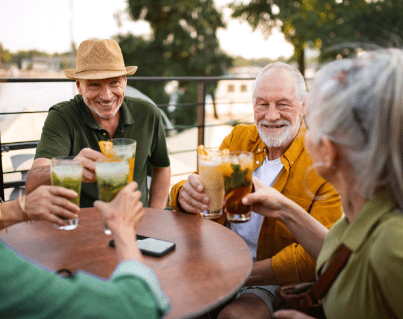 Four friends smiling and clinking iced cocktails around a round outdoor table at a sunny riverside café.