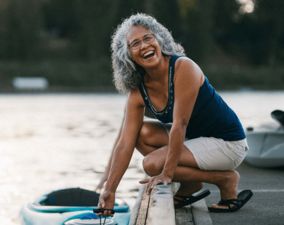Person with curly gray hair and glasses crouching on a dock, smiling while securing a kayak beside the water.